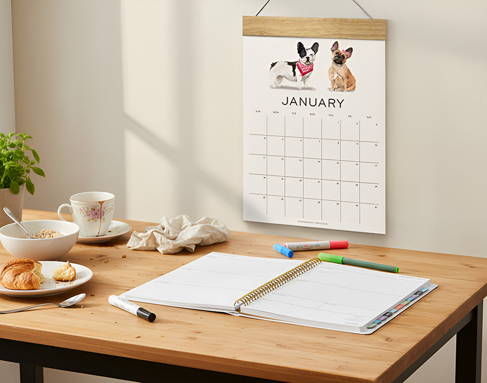 Open weekly planner on a cozy breakfast table with tea, pastries, and markers, with a dog-illustrated January wall calendar hanging above in natural morning light.