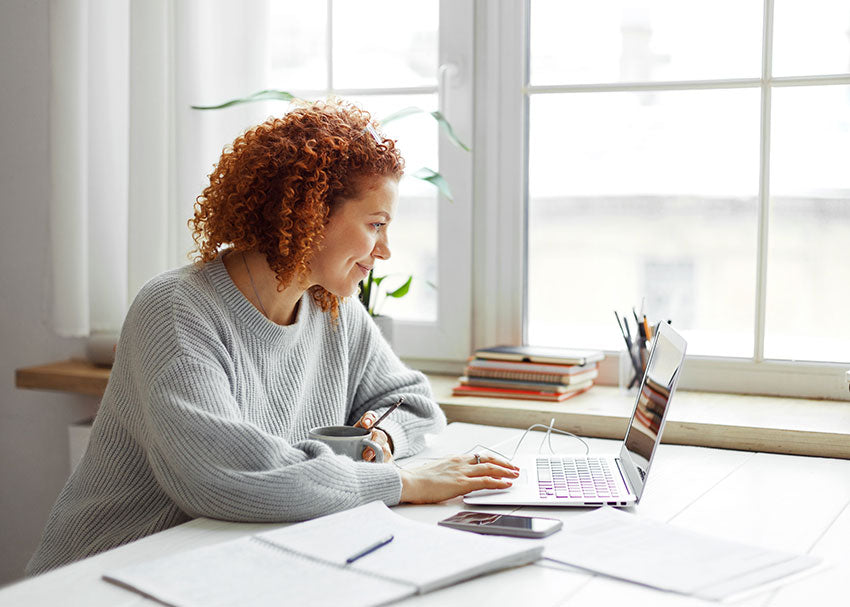 Teacher planning lessons at desk near window with notebook, papers, and laptop in natural daylight