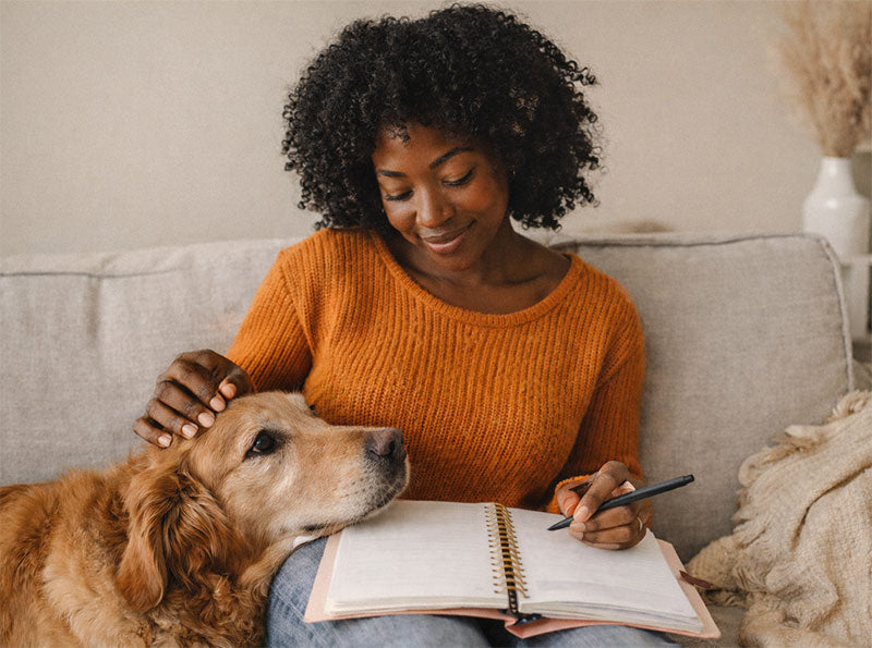 Young woman sitting on a cozy sofa, writing in her planner with a pen while gently petting her golden retriever, surrounded by warm lighting and a relaxed home.