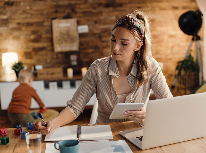 New mom multitasking at home with a planner, tablet, and laptop while toddler plays in the background – staying organized during early motherhood.