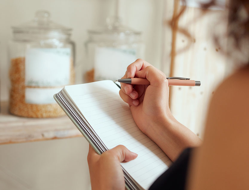 Close-up of a woman writing in a lined notebook while standing in a kitchen, with pantry jars filled with dry goods in the background.