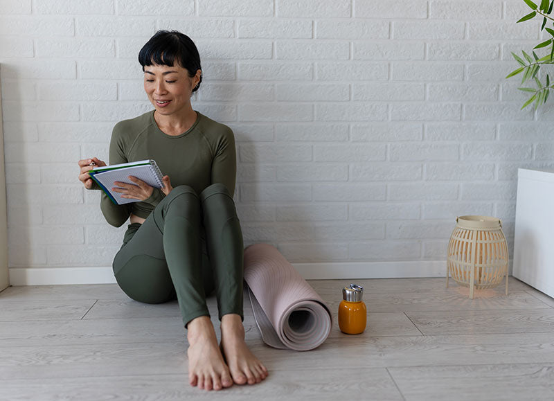 Woman in activewear journaling with a planner beside a yoga mat, juice, and candle — creating a calm, mindful moment after a wellness routine.