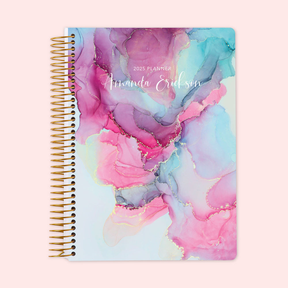 Weekly planner featuring a stunning pink-and-blue abstract ink cover resembling fluid watercolor art, paired with a robust spiral binding and custom name personalization for durable, organized planning with artistic flair.