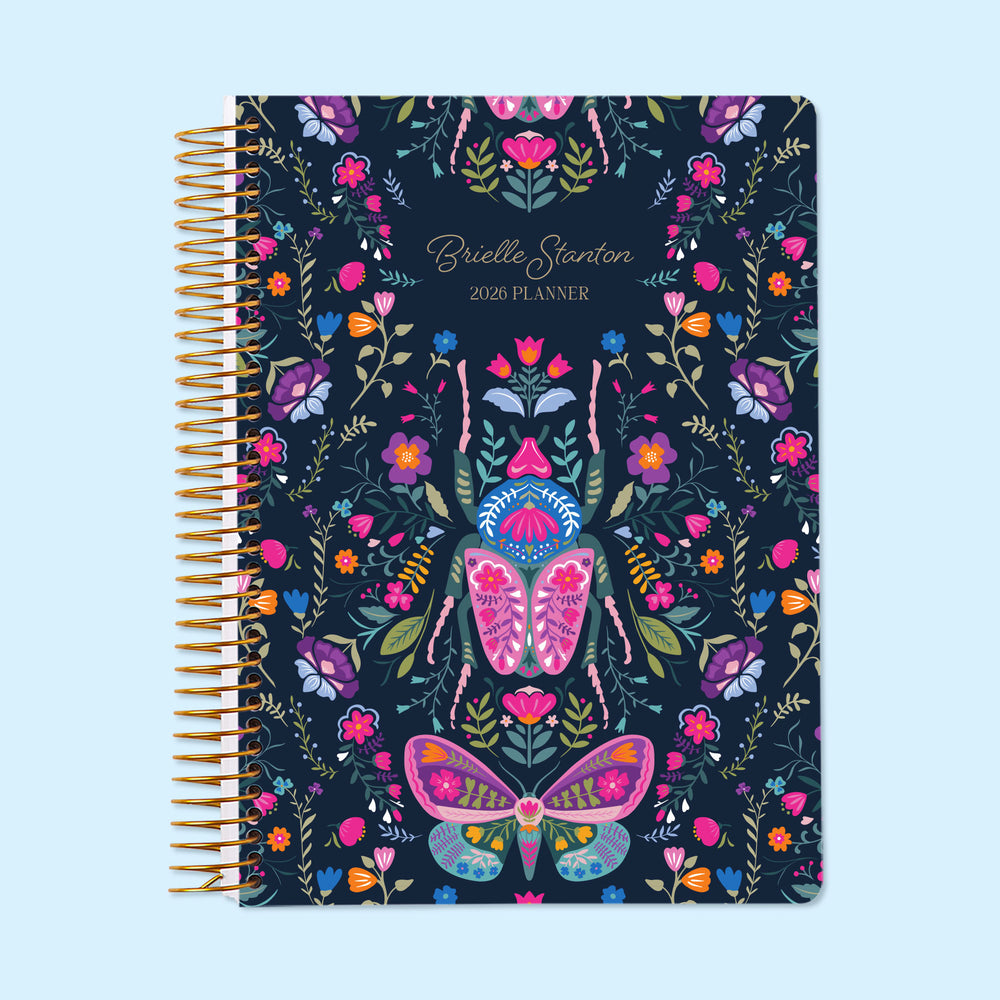6×9 Midnight Garden Wings personalized monthly planner with customizable start month, gold coil binding, and a deep navy floral cover featuring illustrated butterflies, moths, and vibrant garden botanicals.